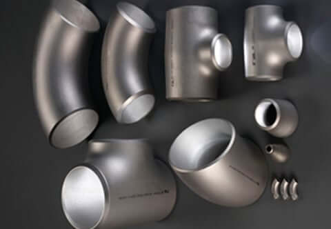 A collection of metallic pipe fittings in various shapes and sizes displayed on a dark surface, including elbows, tees, reducers, and couplings, all appearing to be made from stainless steel.